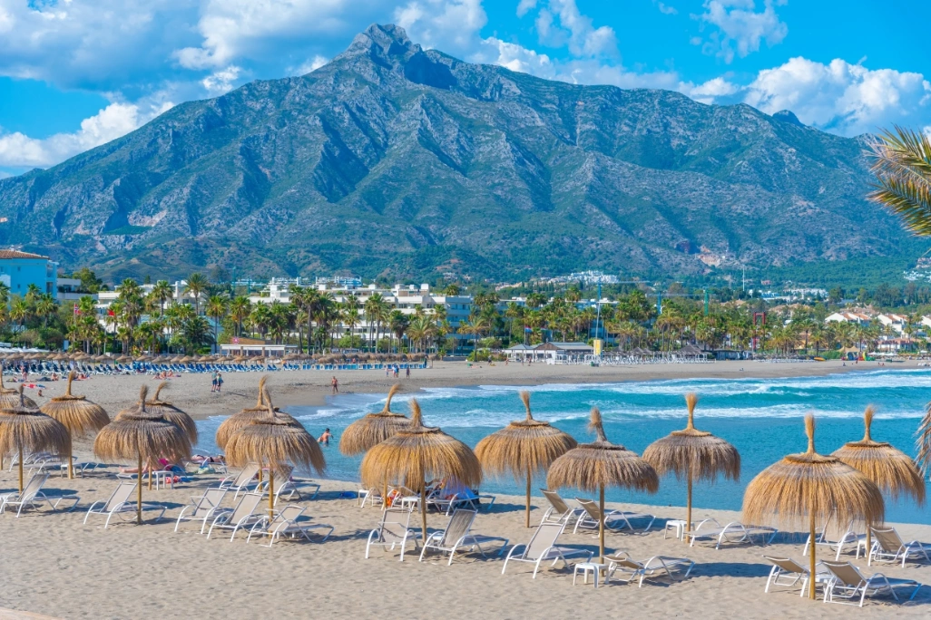 Playa de Puerto Banús, en Marbella, con sombrillas de paja, arena, mar y la montaña de La Concha al fondo