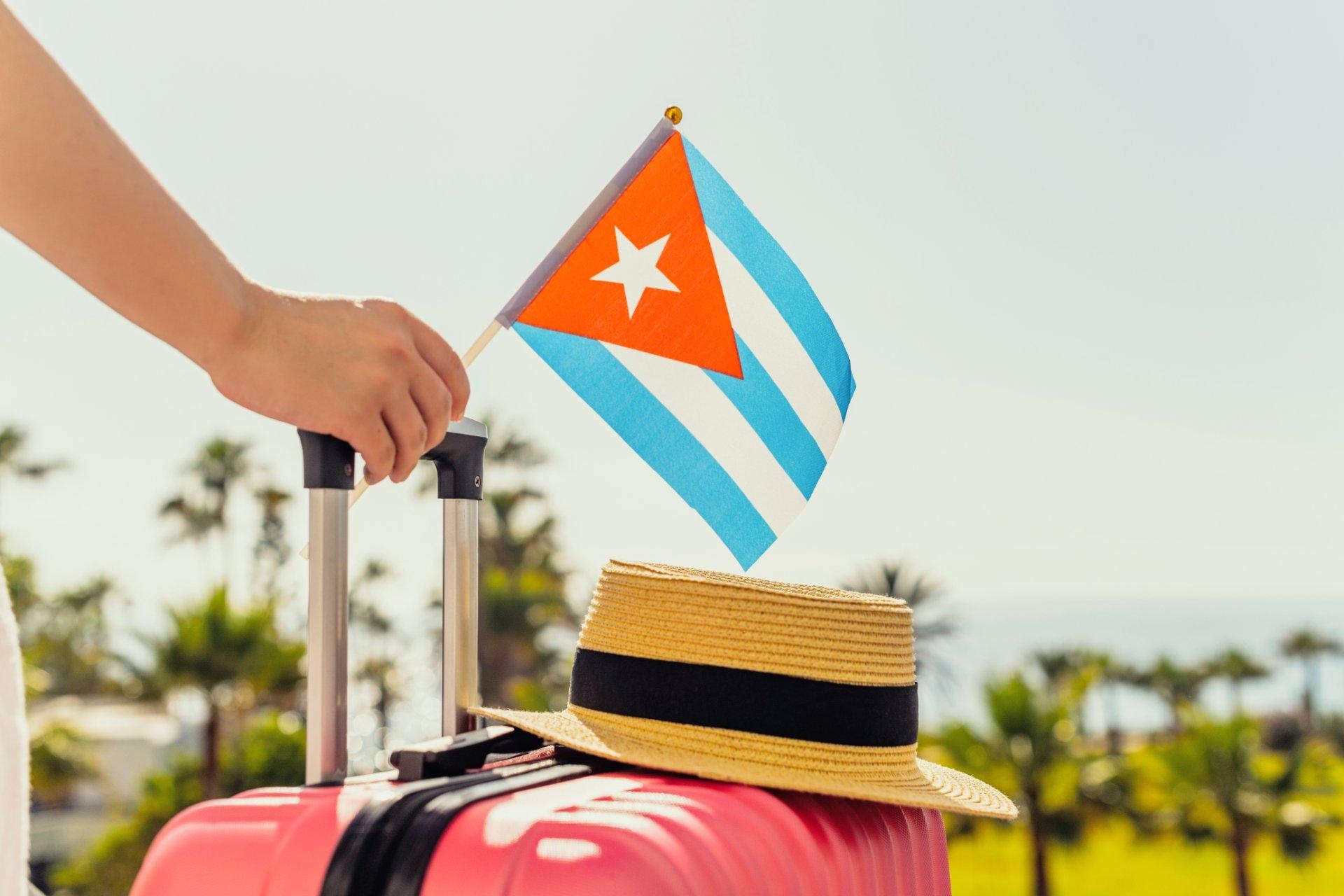 Mujer con maleta rosa, sombrero y bandera cubana saliendo del avión frente a la costa marítima con palmeras. 